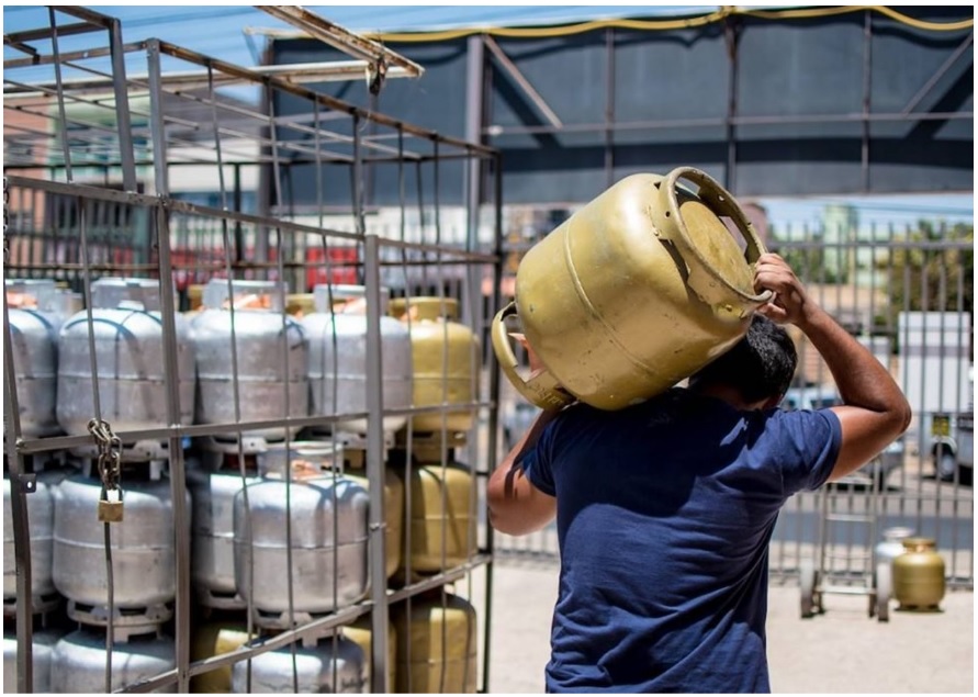 Homem carregando no ombro um botijão de gás no pátio de uma distribuidora de gás