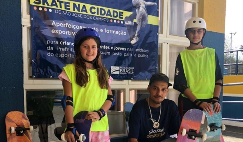 Dois jovens utilizando skates e kits de segurança ao lado de um instrutor do esporte