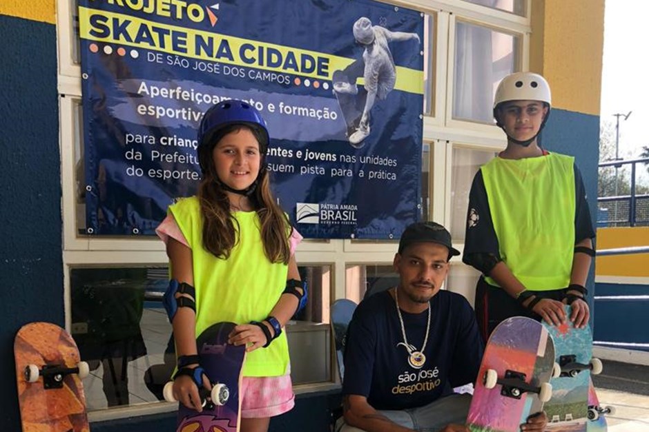 Dois jovens utilizando skates e kits de segurança ao lado de um instrutor do esporte