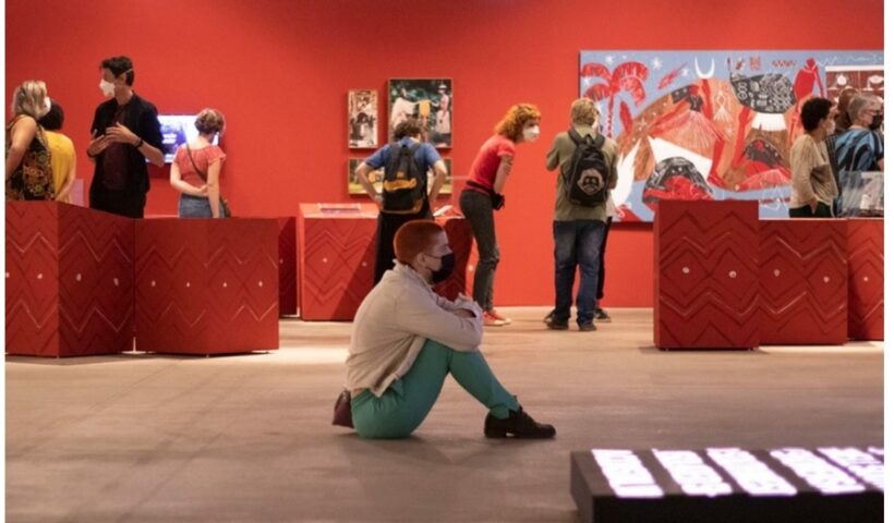 Rapaz sentado no meio de uma exposição de artes, lembra que Campos do Jordão recebe a 34ª exposição itinerante da Bienal de São Paulo
