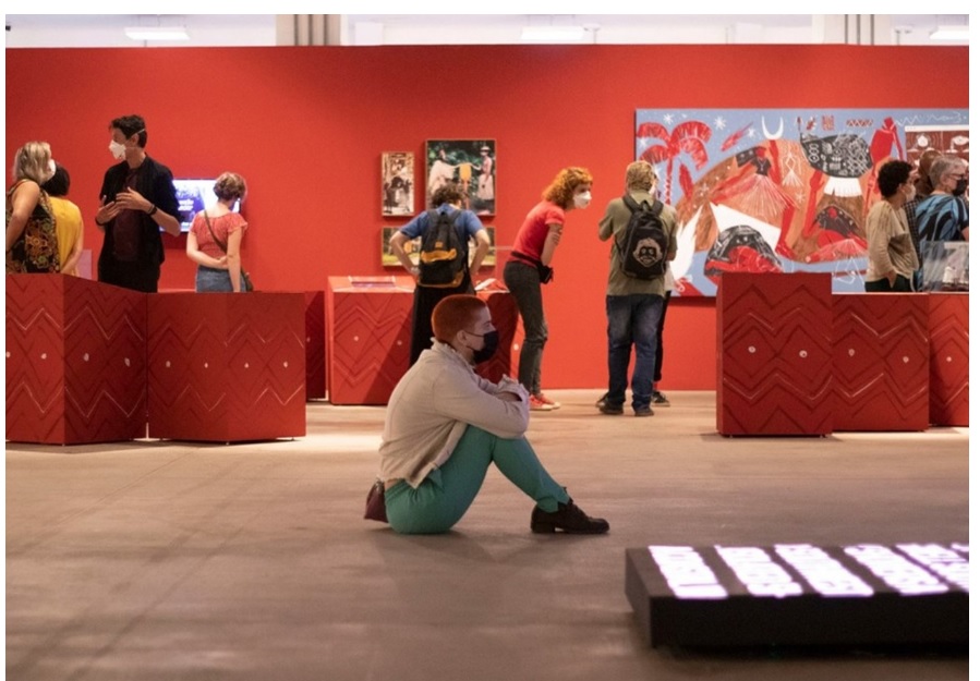 Rapaz sentado no meio de uma exposição de artes, lembra que Campos do Jordão recebe a 34ª exposição itinerante da Bienal de São Paulo