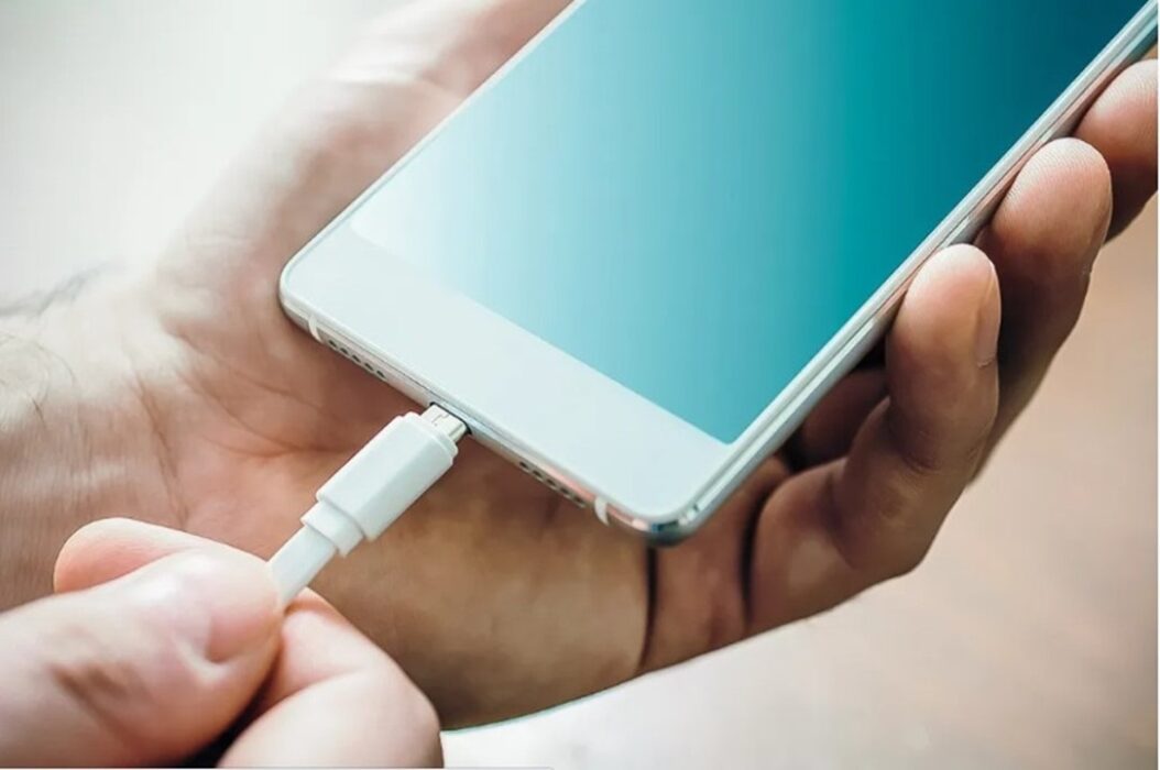 Mãos segurando iPhone e carregador de celular. Venda de iPhone está proibida no brasil sem a inclusão de carregador