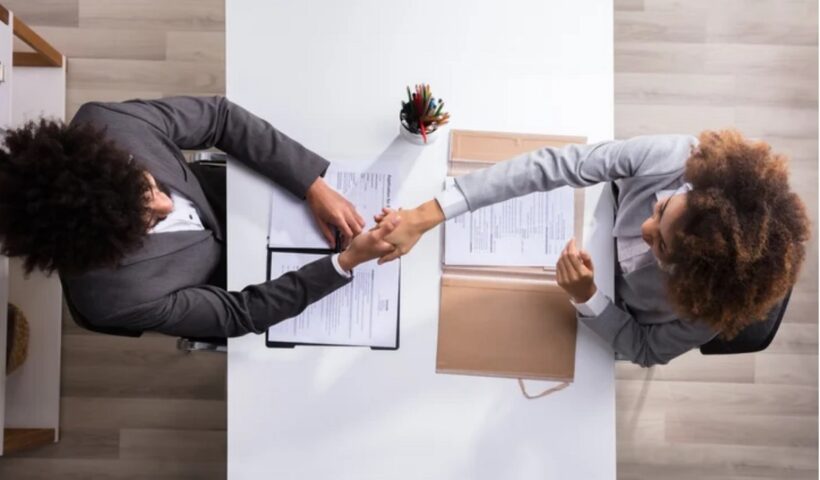 imagem vista de cima de duas mulheres apertando as mão em processo seletivo de emprego