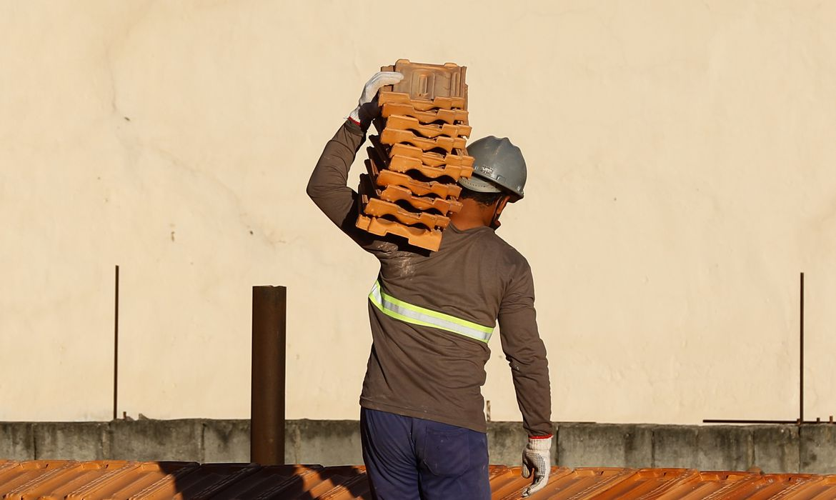 trabalhador da construção civil, com uniforme cinza e azul, luvas, capacete e um bloco de tijolos no ombro. inflação