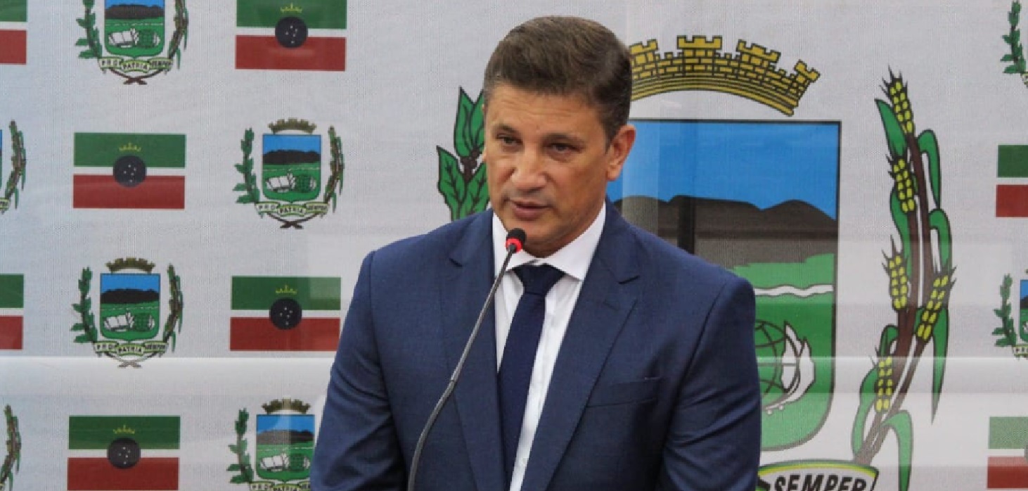 prefeito de pinda vestindo terno azul escuro e gravata fala diante de microfone; símbolo da prefeitura de pindamonhangaba ao fundo