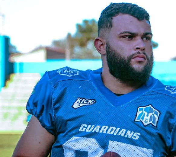 baleado em cruzeiro, Pellizer aparece de uniforme de futebol americano azul, olhando para a direita, no campo de futebol com grama verde