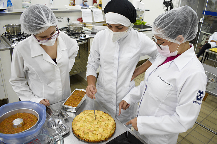 tres alunas do curso de ferias da unitau, vestindo avental branco, cortam fatia de torta preparada em curso