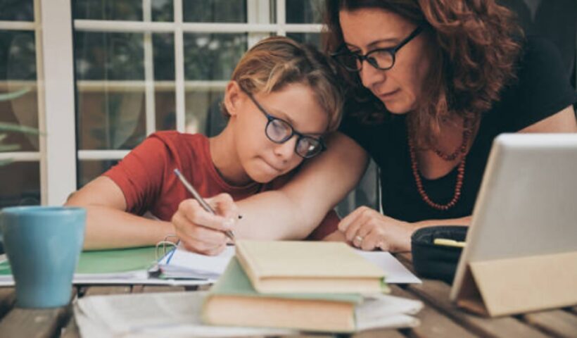 mulher de oculos e cabelo ondulado acompanha menina loira, tambem de oculos, nos estudos durante ensino domiciliar; há livros e cadernos sobre a mesa de casa.