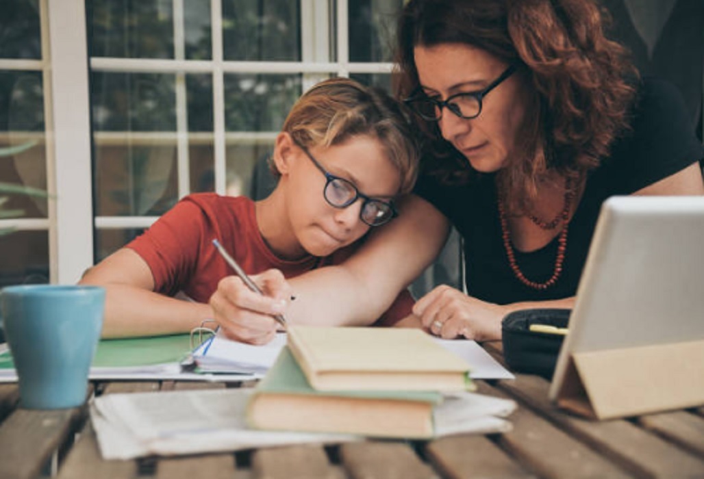 mulher de oculos e cabelo ondulado acompanha menina loira, tambem de oculos, nos estudos durante ensino domiciliar; há livros e cadernos sobre a mesa de casa.
