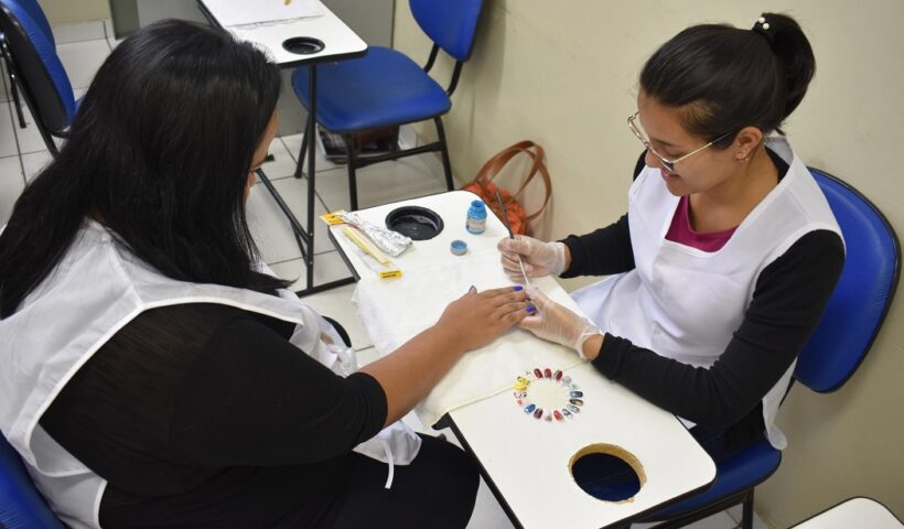 manicure de oculos e avental branco faz as unhas de cliente vestida de preto; escola do trabalho de taubate oferece cursos profissionalizantes gratuitos