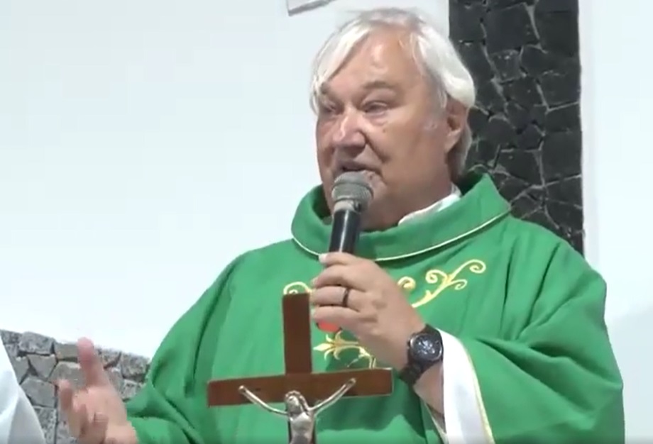 frei hans usa veste religiosa de cor verde, fala ao microfone diante de um crucifixo de madeira