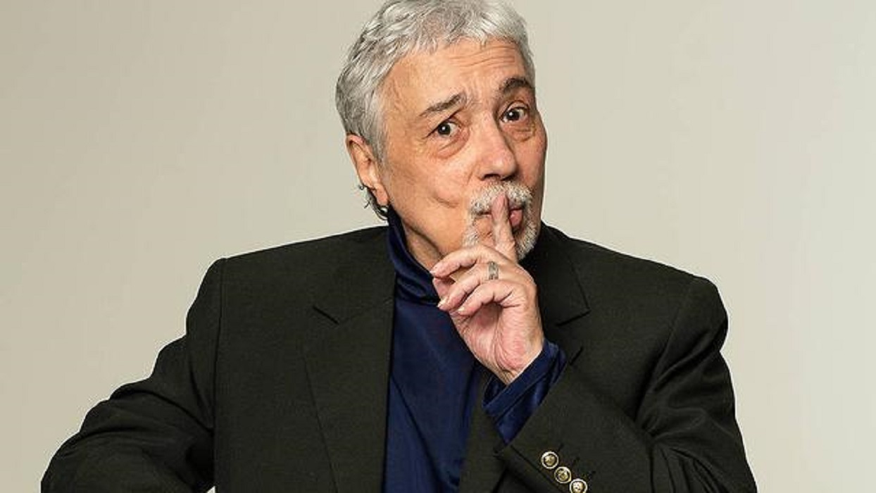 ator pedro paulo rangel aparece na imagem com terno preto, camisa azul escuro e faz sinal de silencio com a mao esquerda