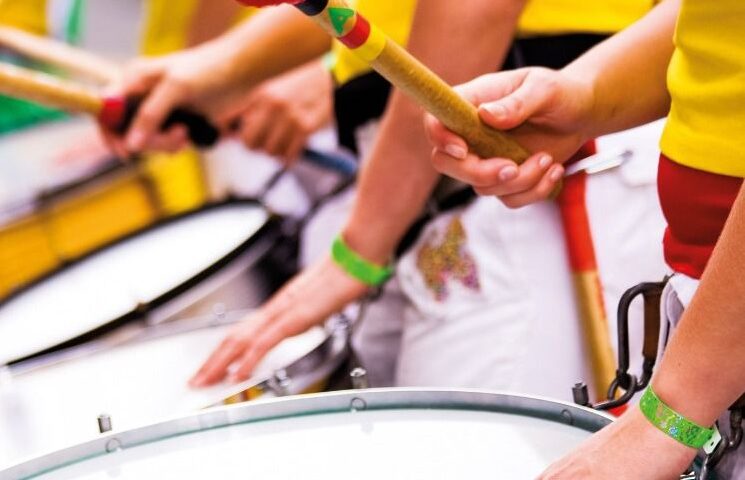 Imagem do projeto plantando samba mostra mãos tocando tambores; as pessoas vestem camisetas amarelas e têm fitas verdes amarradas nos pulsos