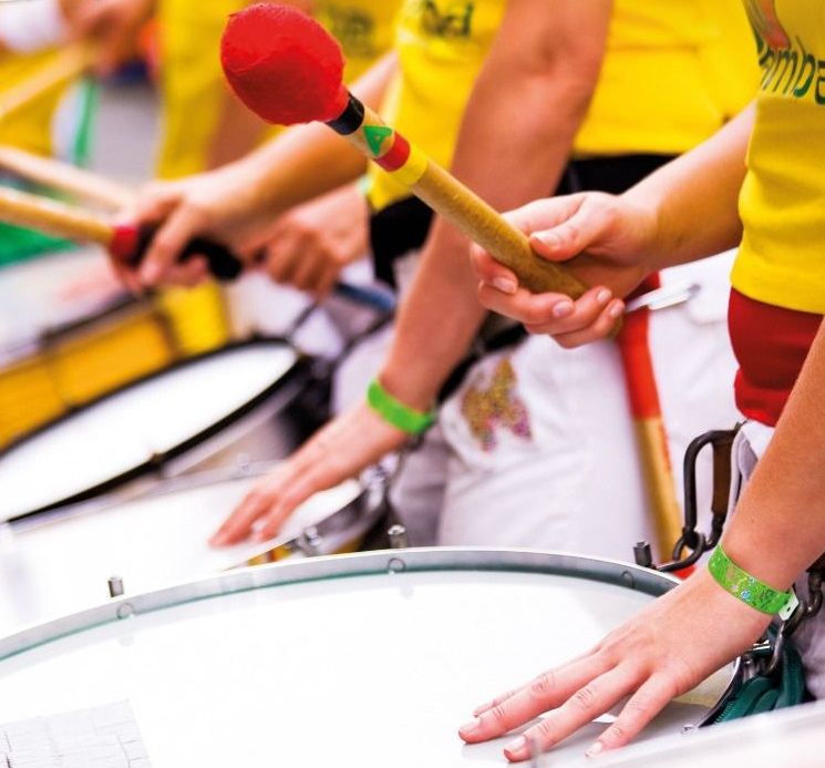 Imagem do projeto plantando samba mostra mãos tocando tambores; as pessoas vestem camisetas amarelas e têm fitas verdes amarradas nos pulsos
