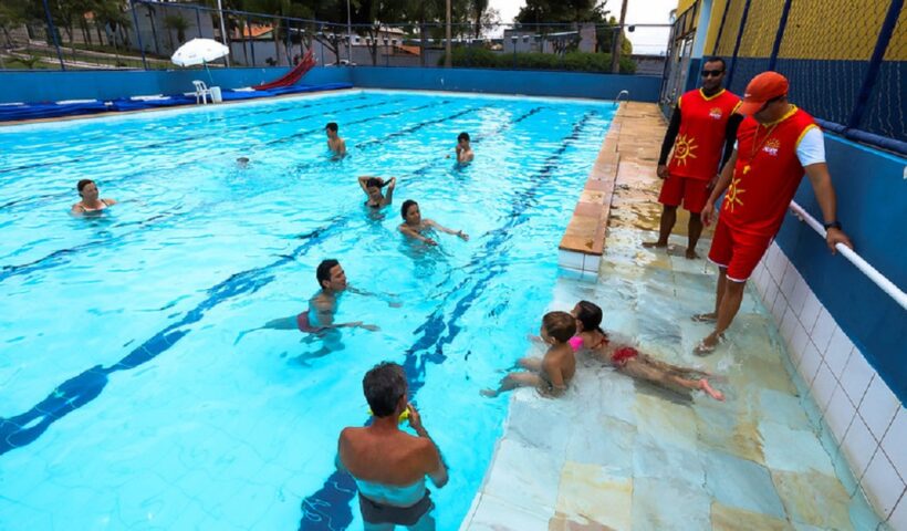 piscina do projeto verao de sao jose dos campos, com sete pessoas dentro, entre adultos e crianças, e dois guarda-vidas, com uniforme vermelho, do lado de fora