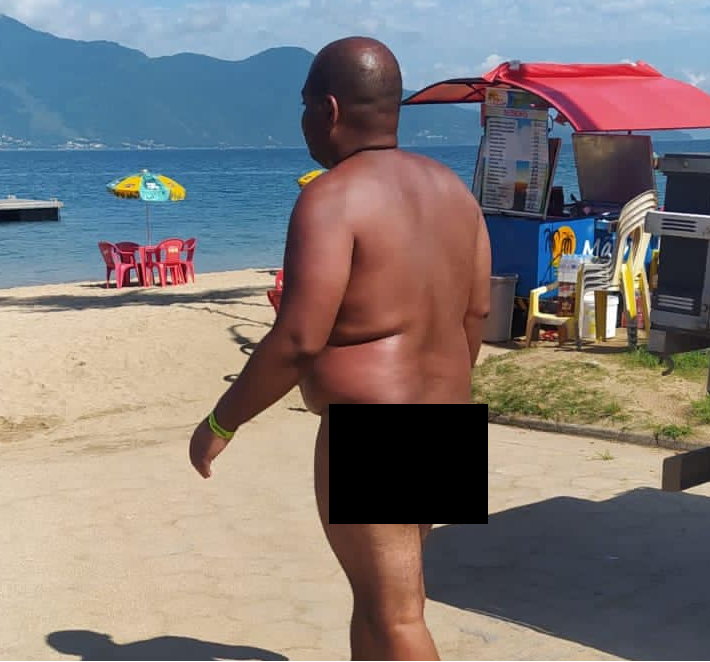 homem totalmente despido caminha na areia da praia de Ilhabela; há barracas em volta. ele está acima do peso e tem a pele negra e bronzeada