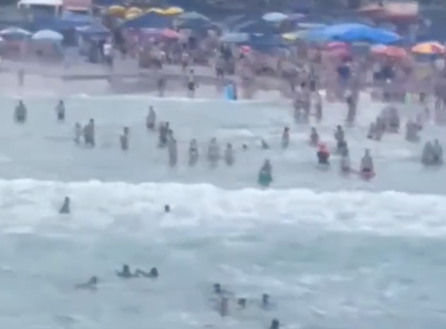 Helicóptero Águia resgata 4 de afogamento em Ubatuba; duas eram cegas; mar com ondas, muitos turistas na faixa de areia, várias pessoas dentro do mar, entre elas as que se afogam