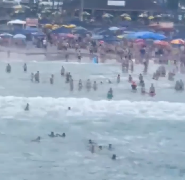 Helicóptero Águia resgata 4 de afogamento em Ubatuba; duas eram cegas; mar com ondas, muitos turistas na faixa de areia, várias pessoas dentro do mar, entre elas as que se afogam