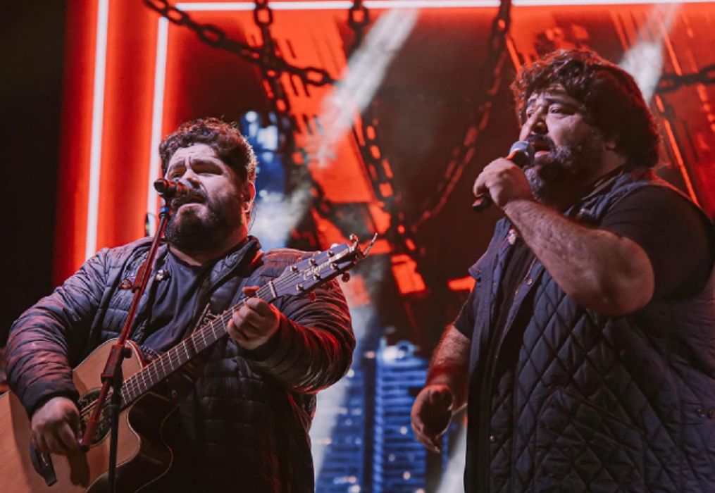 atrações no vale incluem cesar menotti e fabiano e carnaval; dupla sertaneja canta sobre o palco, segurando violão, em frente ao microfone