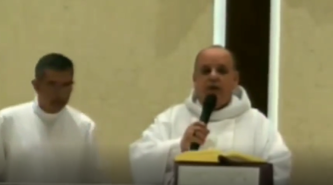 padre antonio, com roupa religiosa branca, biblia aberta sobre o púlpito, fala ao microfone; ele foi chamado de comunista na missa