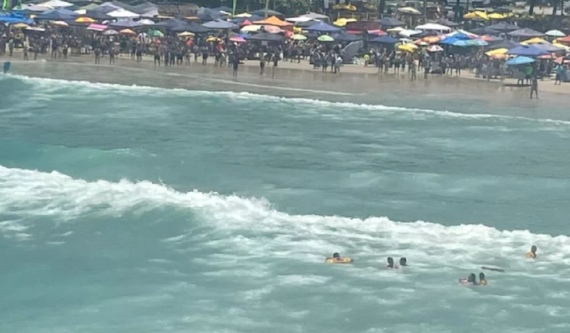 mar com ondas e trio se afogando em corrente maritima; a faixa de areia está repleta de turistas e guarda-sois