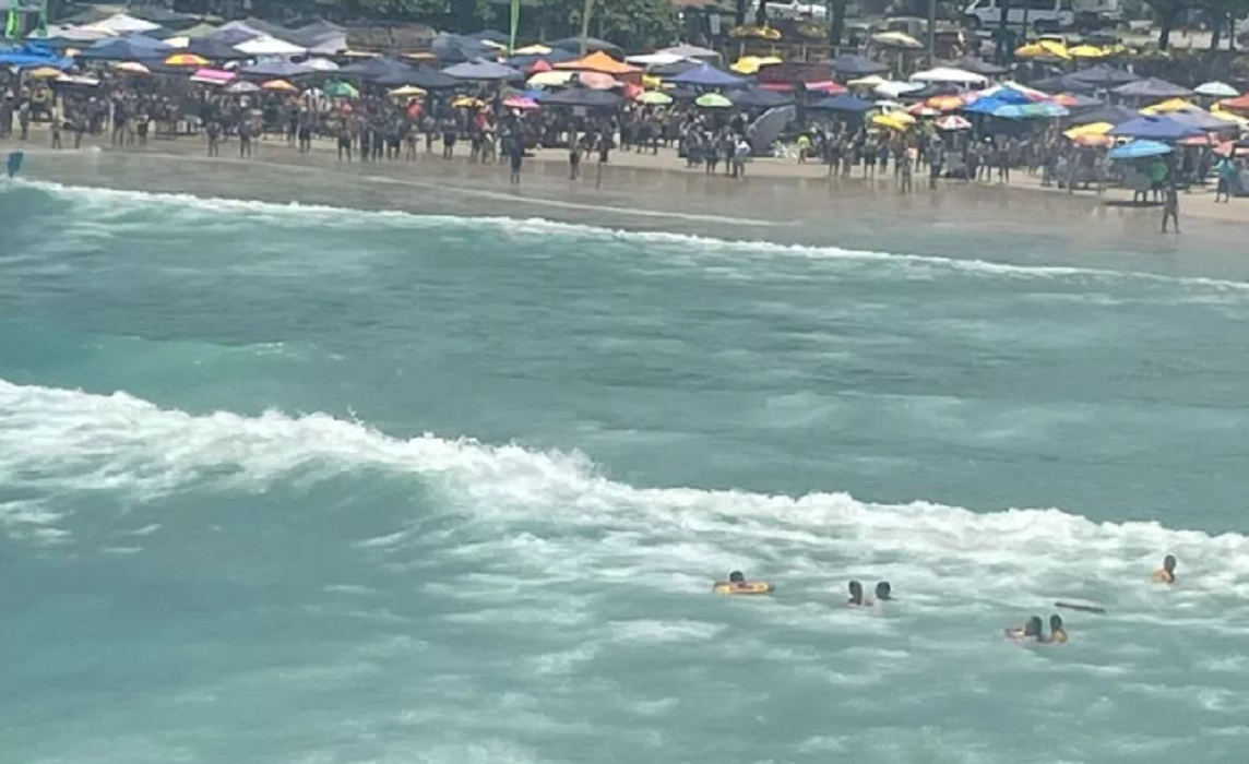 mar com ondas e trio se afogando em corrente maritima; a faixa de areia está repleta de turistas e guarda-sois