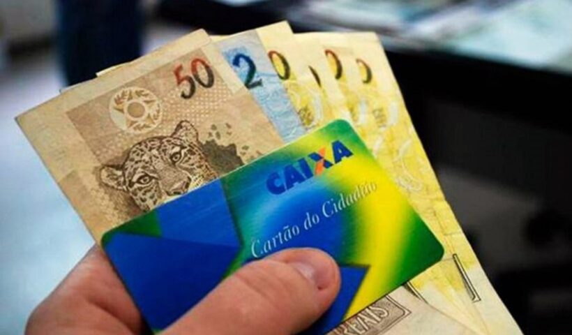 Mão segurando cartão da Caixa e dinheiro. Pagamento de abono Pis/pasep começa a partir de abril