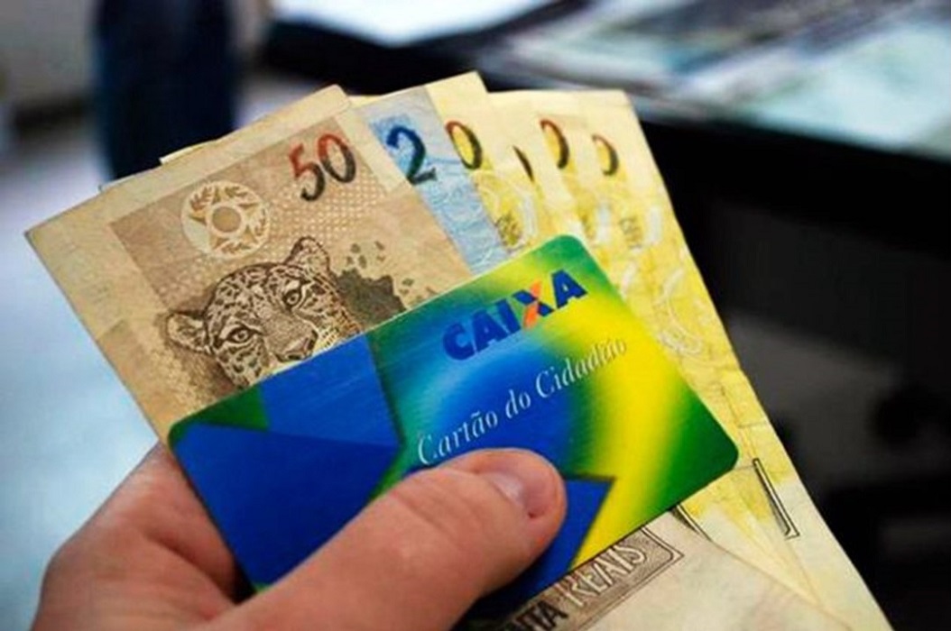 Mão segurando cartão da Caixa e dinheiro. Pagamento de abono Pis/pasep começa a partir de abril