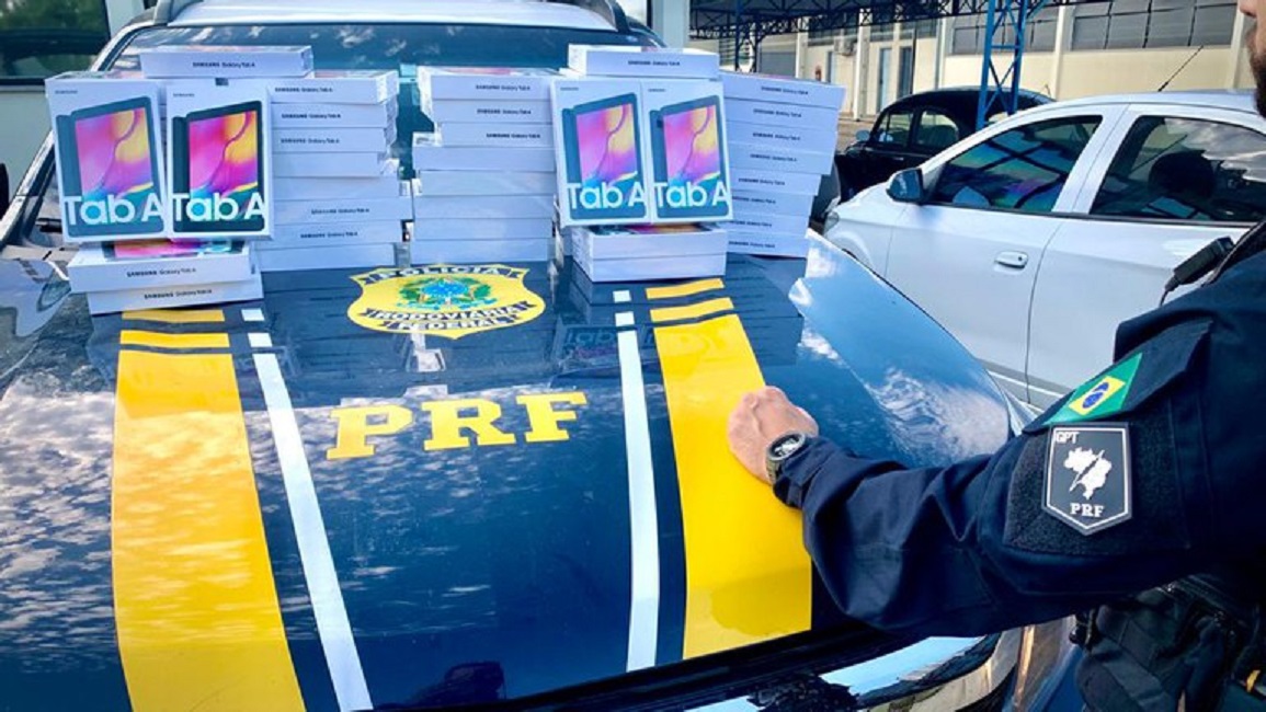 Policial rodoviário apontando para Tablets sobre o capô de viatura da PRF. PRF apreende carga de eletrônicos sem comprovação Fiscal na Via Dutra, em Taubaté