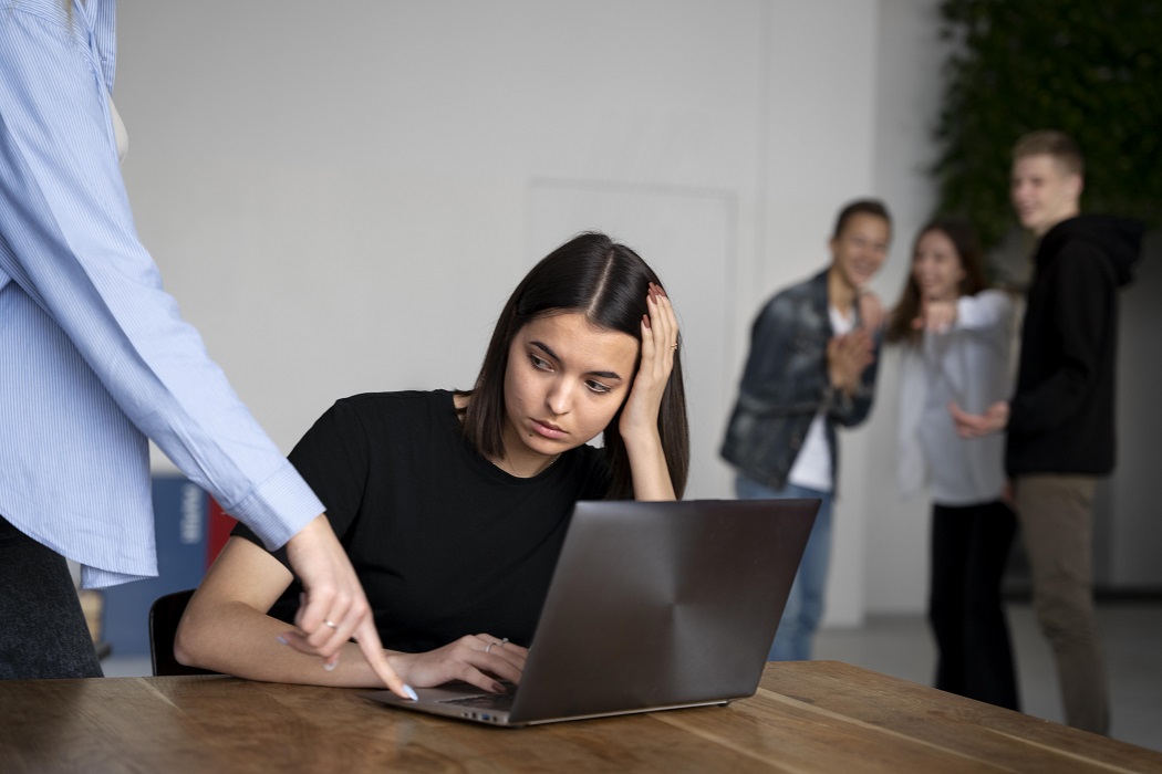 Mulher em frente ao computador recebe crítica de superior. Pessoas ao lado zombam da funcionária. Tema: assédio moral