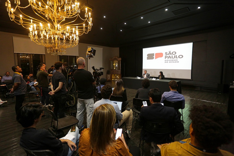 Governador Tarcísio de Freitas sentado junto á uma mesa em entrevista coletiva com repórteres.