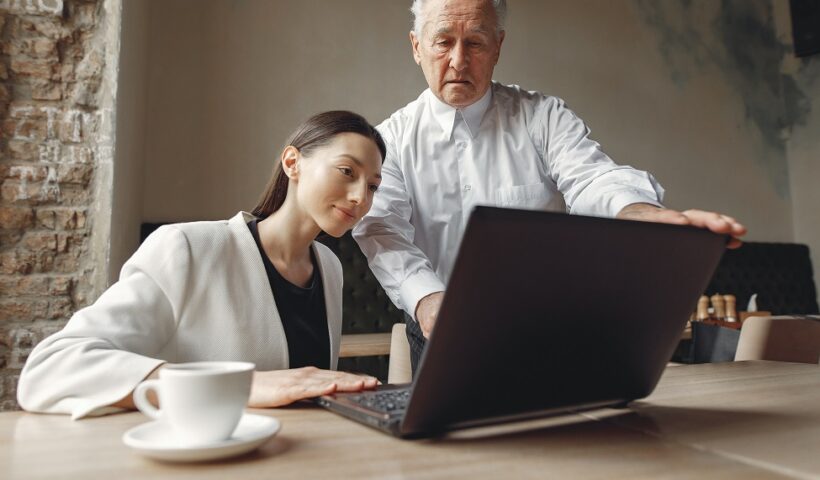 Mulher jovem olha para computador ao lado de homem idoso que parece dar orientações de trabalho. CBN Mercado de Trabalho: Gerações no Mercado de Trabalho e seus desafios
