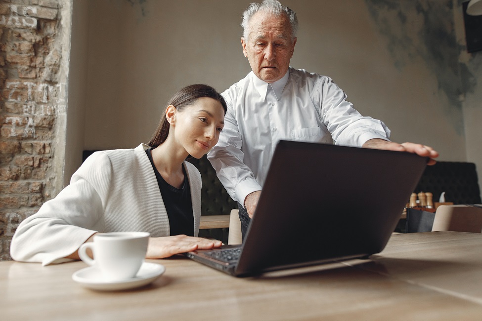 Mulher jovem olha para computador ao lado de homem idoso que parece dar orientações de trabalho. CBN Mercado de Trabalho: Gerações no Mercado de Trabalho e seus desafios