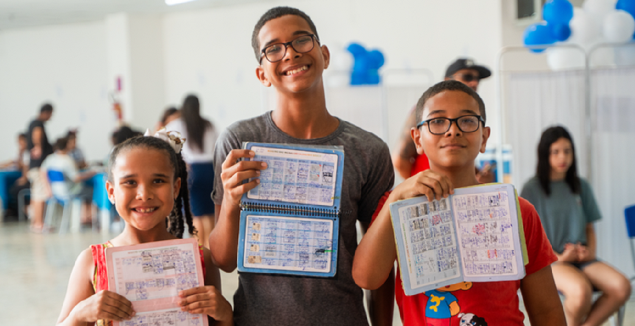 Três crianças sorrindo mostras suas cardenetas de vacinação completas. Vacina contra Covid-19 será incluída no calendário nacional de crianças e grupos prioritários a partir de 2024