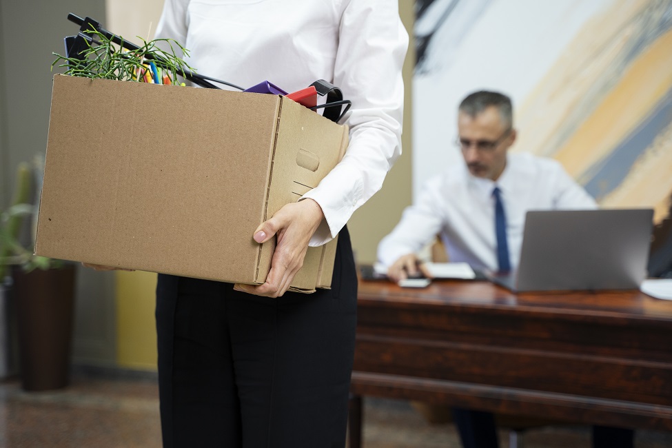 mulher carregando em vista lateral, uma caixa de papelão com seu chefe ao fundo. CBN Mercado de Trabalho: Fui demitido, e agora? Acalma o coração
