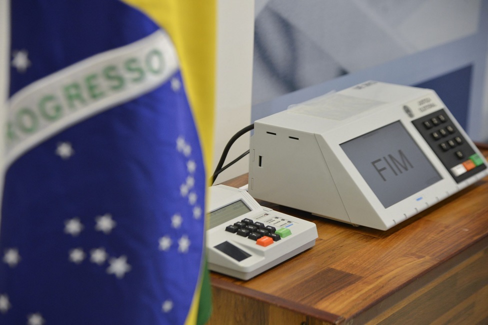 urna eletrônica de votação em cima de uma mesa, e em primeiro plano, a bandeira do Brasil. TSE inicia teste público das urnas eletrônicas para eleições de 2024
