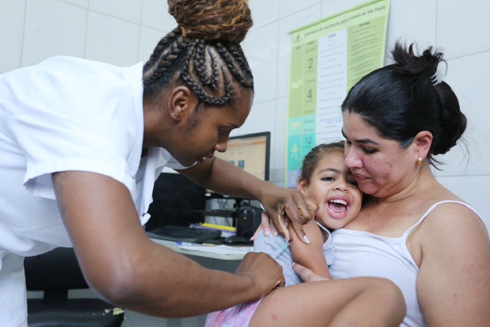 mãe segurando filha que está sendo vacinada por agente de saúde. Calendário nacional da vacinação contra a covid inclui as crianças