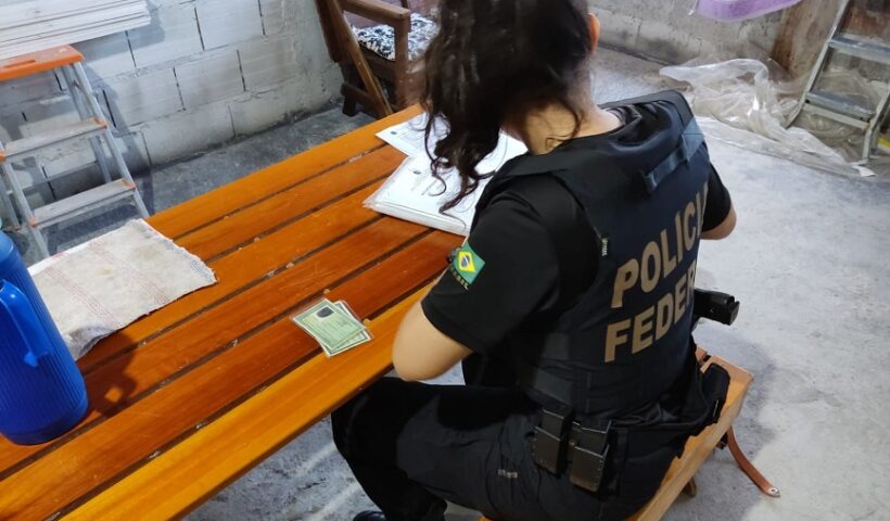Agente mulher da PF de costas, sentada de frente a uma mesa com documentos. Homem é preso por se passar por menina e extorquir fotos íntimas de menores, em Jacareí