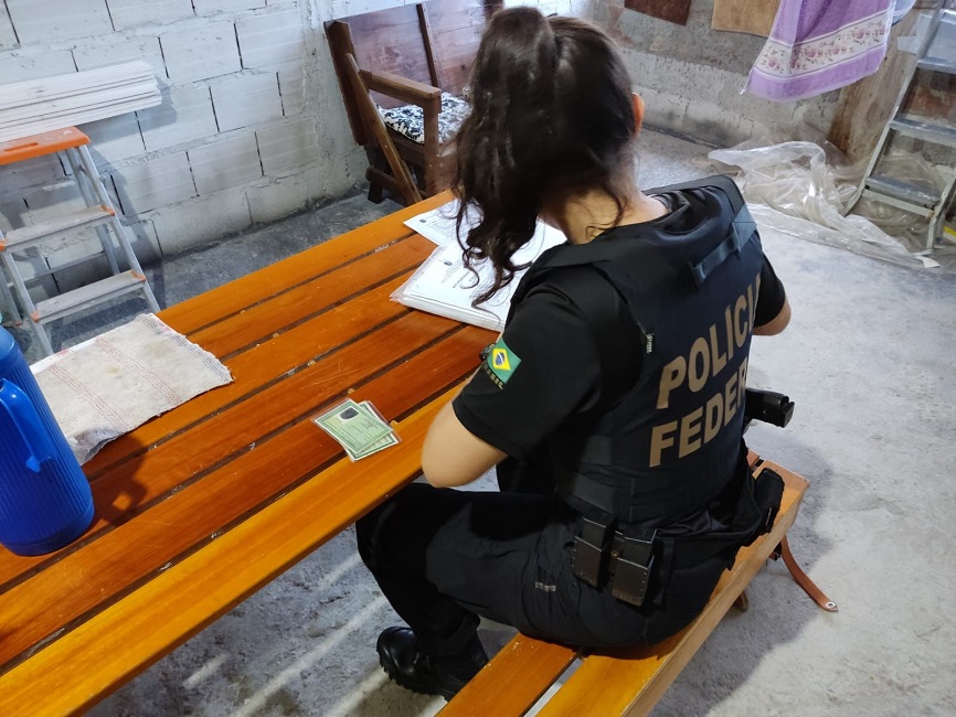 Agente mulher da PF de costas, sentada de frente a uma mesa com documentos. Homem é preso por se passar por menina e extorquir fotos íntimas de menores, em Jacareí