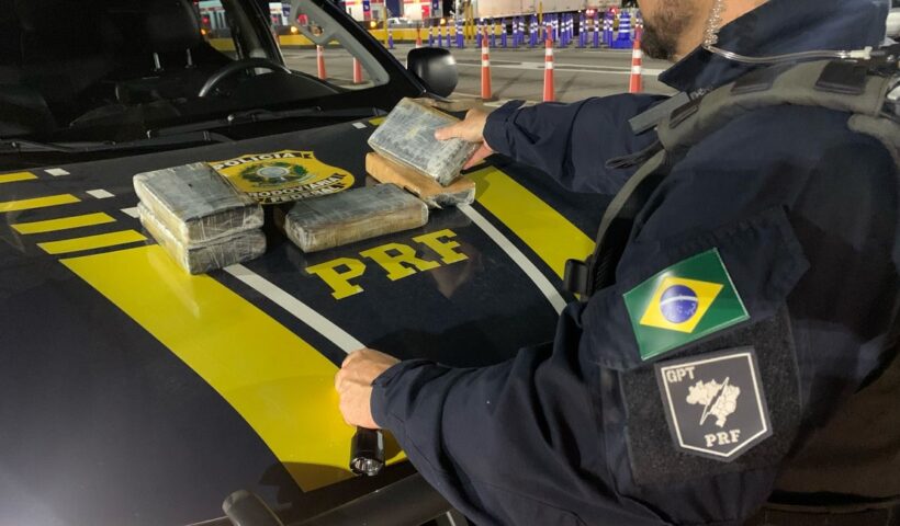 Perfil de agente da PF colocando pacotes de drogas no capô de viatura.