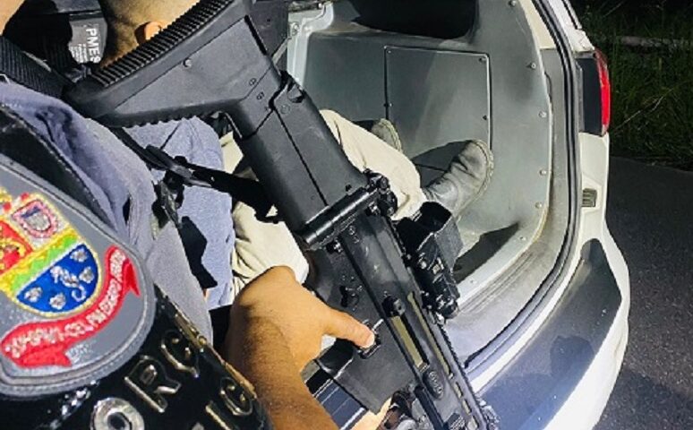 PM com fuzil acompanha suspeito algemado sair de viatura policial. Operações da PM resultam em prisões por diversos crimes no litoral norte de São Paulo