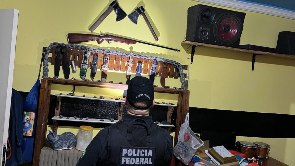 Policial federal de costas olha parede com armas expostas. PF prende homem com tabela de preços de vídeos contendo exploração sexual infantojuvenil, em Taubaté