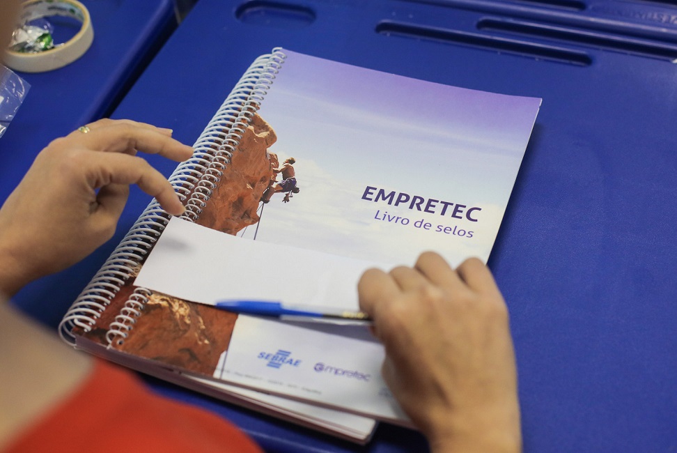 Mão segurando uma caneta azul, em cima de uma apostila, apoiada sob uma mesa azul, ilustrando que o Sebrae abriu inscrições para o curso Empretec em São José dos Campos.