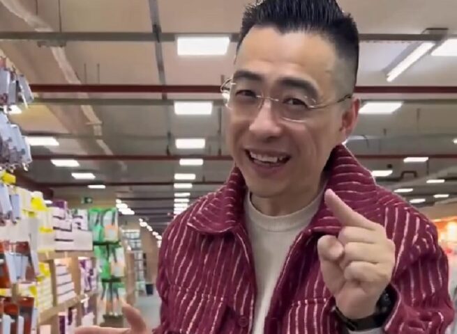 Alex Ye, criador de conteúdo de ascendência chinesa, aparece sorridente em um corredor de loja vestindo uma jaqueta de veludo bordô enquanto faz gestos explicativos a câmera.