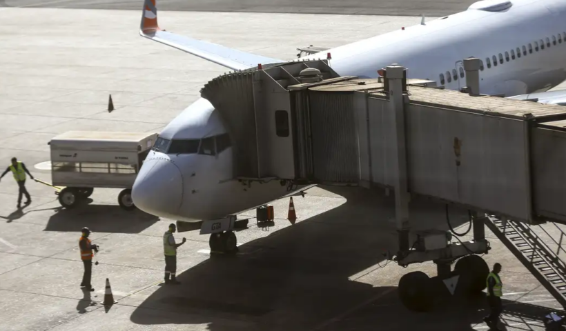 Um avião comercial de passageiros está acoplado a uma ponte de embarque em um pátio de aeroporto iluminado pelo sol, enquanto funcionários de solo realizam procedimentos de serviço ao redor da aeronave