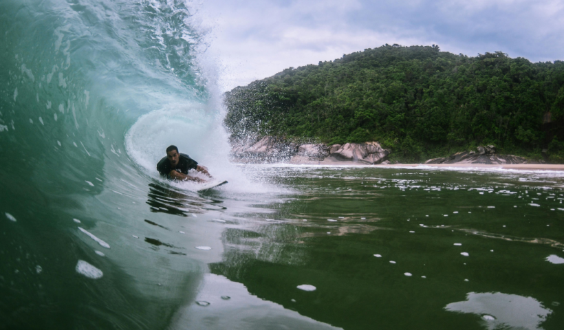 Um surfista executa uma manobra ágil dentro de um tubo de uma onda cristalina, com uma encosta de vegetação tropical densa ao fundo.
