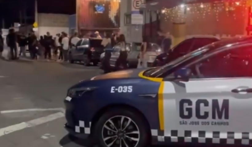 Carro da GCM em uma rua com diversas pessoas na calçada, é noite e as luzes do carro estão ligadas