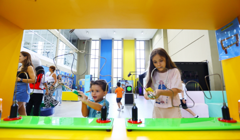 Duas crianças interagem com brinquedos educativos de metal em um museu de ciências, emolduradas por uma estrutura amarela em primeiro plano