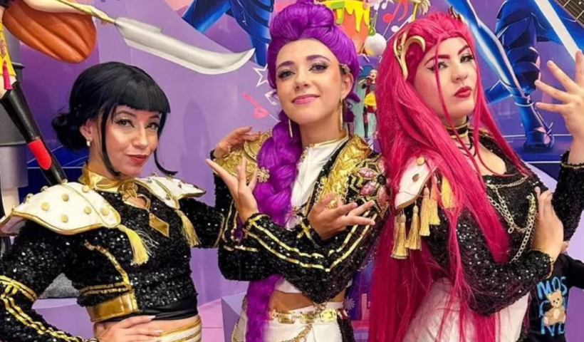 Três mulheres vestidas como personagens do filme Guerreiras do K-pop, da Netflix, posam juntas usando figurinos coordenados em preto e dourado, com perucas estilizadas nas cores preta, roxa e rosa vibrante