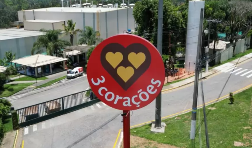 Fachada da fábrica da 3 corações, com um prédio ao fundo e um outdoor mostrando o logo da 3 corações em vermelho
